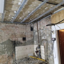 LUK-BUD Łukasz Wołoch - Wnętrze w trakcie remontu: surowy ceglany mur częściowo pokryty zaprawą, metalowa konstrukcja podwieszanego sufitu, fragment starej instalacji wodnej i resztki gruzu na piecu kaflowym.