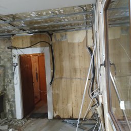 LUK-BUD Łukasz Wołoch - Wnętrze w trakcie remontu: widoczne fragmenty płyty karton gips na suficie, drewniana boazeria na ścianach z resztkami tapety, odsłonięte przewody elektryczne, metalowe profile, gruz na podłodze...