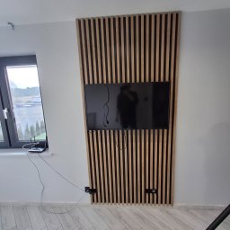 LUK-BUD Łukasz Wołoch - Telewizor zawieszony na ścianie z dekoracyjnych, drewnianych listew w pionowym układzie, widok z boku na jasną podłogę z wzorem jodełki, okno z widokiem na zewnątrz i router na parapecie.