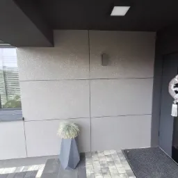 Fragment elewacji budynku z szarymi panelami o delikatnej fakturze, oświetlony lampą elewacyjną, z widocznym fragmentem okna z żaluzjami, dekoracyjnym wieńcem na drzwiach i nowoczesną donicą...