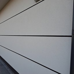 LUK-BUD Łukasz Wołoch - Elewacja budynku z jasnoszarym tynkiem strukturalnym, podzielona czarnymi, poziomymi liniami, z zamontowanym nawiewnikiem wentylacyjnym i rurą spustową.