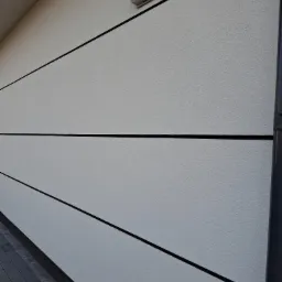 Elewacja budynku z jasnoszarym tynkiem strukturalnym, podzielona czarnymi, poziomymi liniami, z zamontowanym nawiewnikiem wentylacyjnym i rurą spustową.