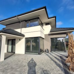 LUK-BUD Łukasz Wołoch - Nowoczesny dom jednorodzinny w Dobre, z elewacją w odcieniach bieli i szarości, widoczne duże okna z żaluzjami, kostka brukowa na podjeździe i ozdobna trawa pampasowa.