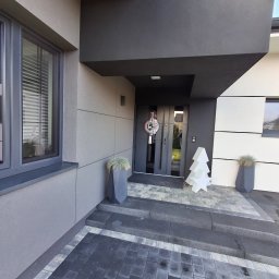 LUK-BUD Łukasz Wołoch - Nowoczesna elewacja domu w odcieniach szarości i bieli, z dekoracyjnym wieńcem na drzwiach wejściowych i podświetlaną choinką na zewnątrz.