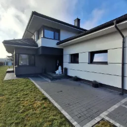 Nowoczesny dom jednorodzinny z biało-szarą elewacją, kostką brukową na podjeździe i minimalistycznymi dekoracjami świątecznymi przed wejściem.