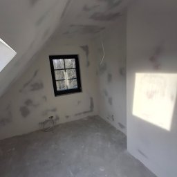 LUK-BUD Łukasz Wołoch - Pokój na poddaszu w trakcie prac wykończeniowych, ściany przygotowane pod malowanie, widoczne ślady po szpachlowaniu, okno z czarną ramą.