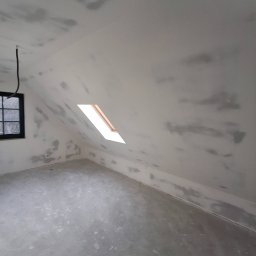 LUK-BUD Łukasz Wołoch - Poddasze w trakcie prac wykończeniowych: surowe ściany i sufit z nałożoną warstwą szpachli, widoczne okno dachowe z drewnianą ramą i czarna rama okienna na bocznej ścianie.