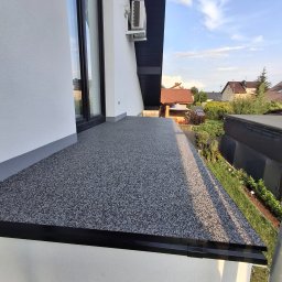 LUK-BUD Łukasz Wołoch - Wykończony balkon z szarą, kamyczkową posadzką i czarnym obramowaniem, widok z góry na tle zieleni i zabudowań. Widoczny fragment elewacji budynku.