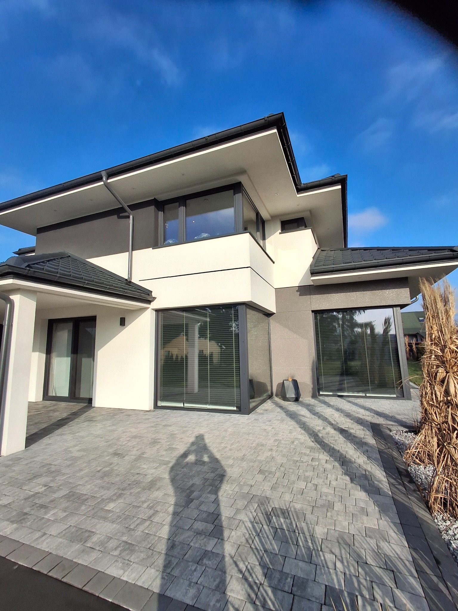 Nowoczesny dom jednorodzinny w Dobre, z elewacją w odcieniach bieli i szarości, widoczne duże okna z żaluzjami, kostka brukowa na podjeździe i ozdobna trawa pampasowa.