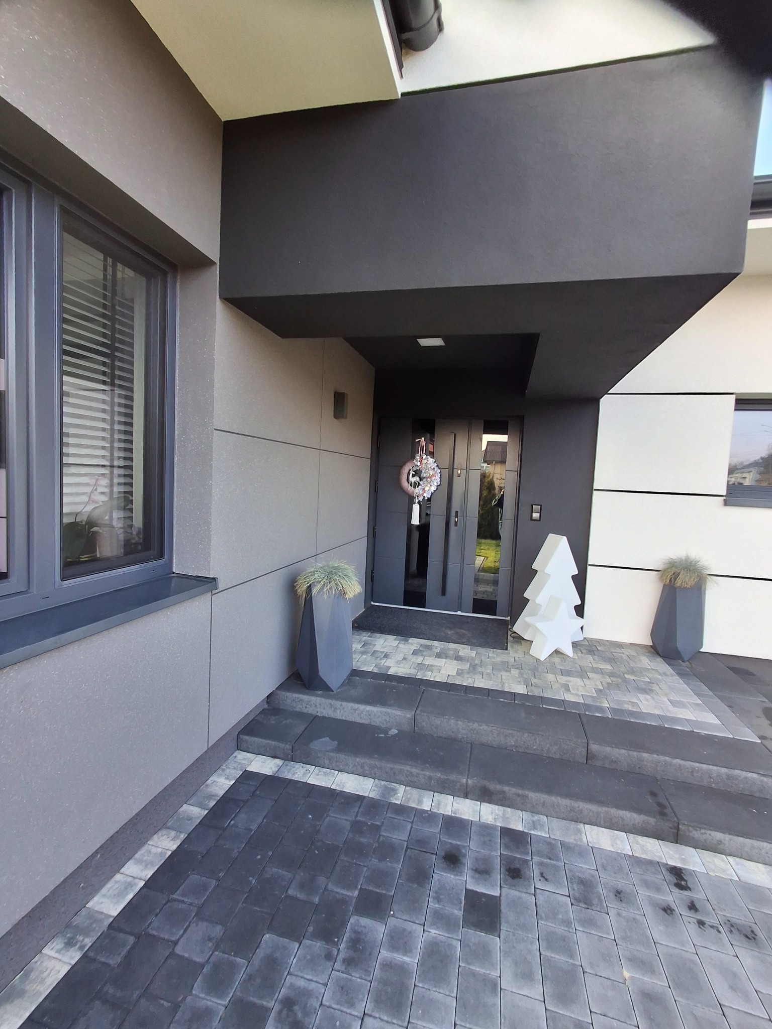 Nowoczesna elewacja domu w odcieniach szarości i bieli, z dekoracyjnym wieńcem na drzwiach wejściowych i podświetlaną choinką na zewnątrz.