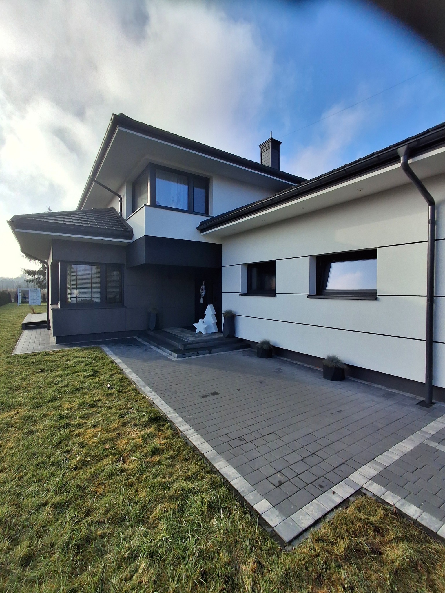 Nowoczesny dom jednorodzinny z biało-szarą elewacją, kostką brukową na podjeździe i minimalistycznymi dekoracjami świątecznymi przed wejściem.