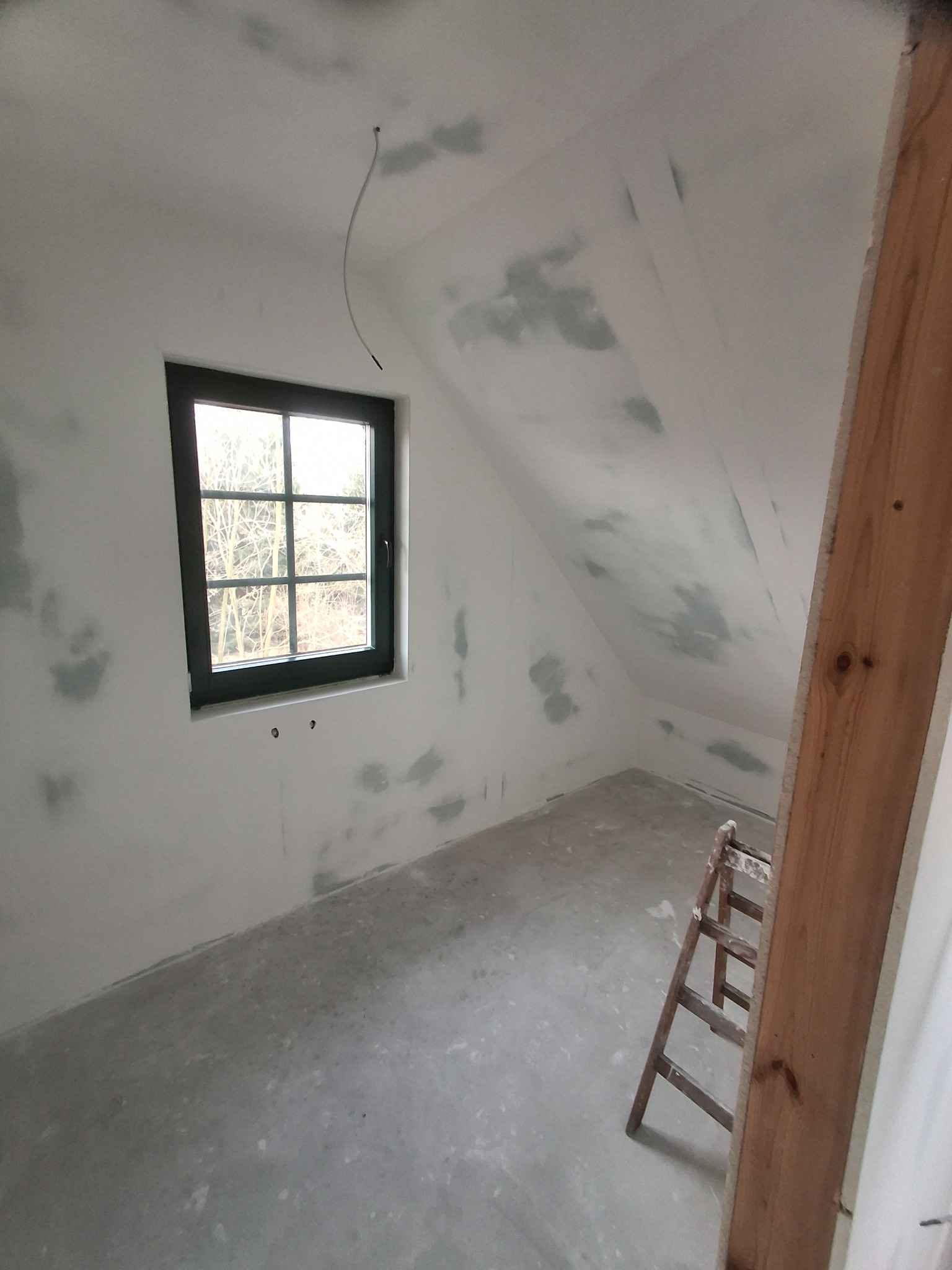 Poddasze w trakcie wygładzania ścian, widoczne ślady po łataniu, okno z ciemną ramą i drewniana drabina oparta o ścianę.