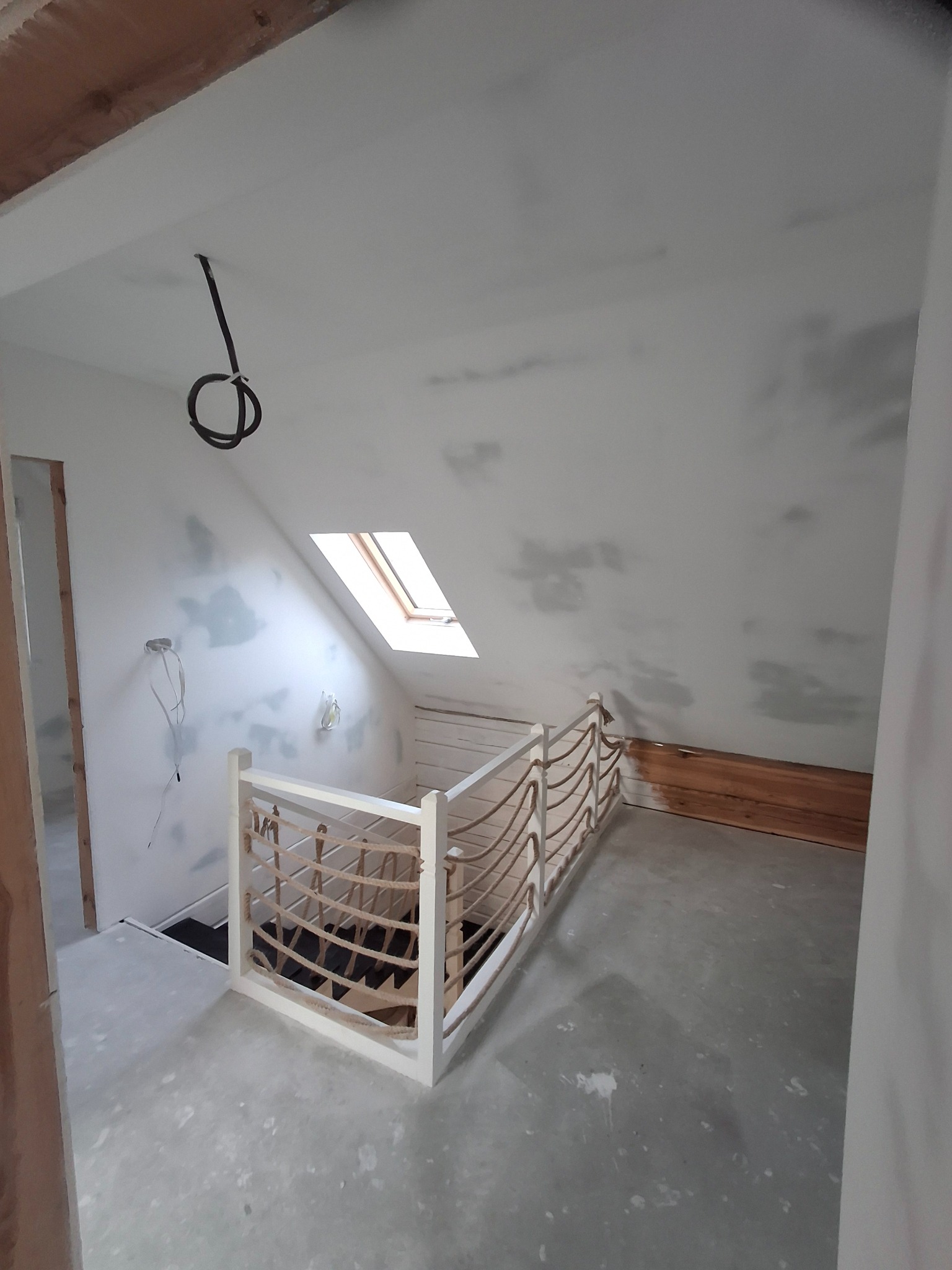 Poddasze w trakcie wykańczania, widoczne szpachlowane ściany, okno dachowe, biała balustrada ze sznurem, wystające kable elektryczne i surowe drewno na podłodze.
