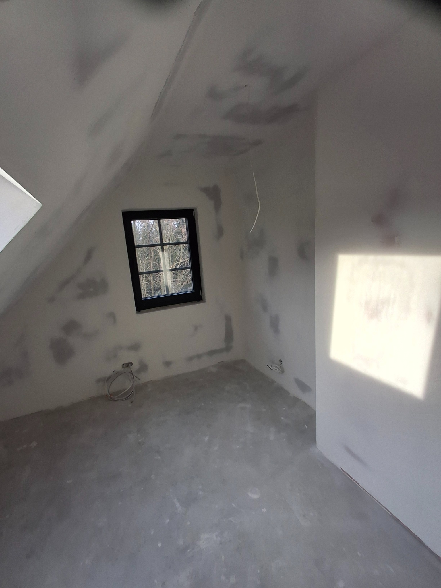 Pokój na poddaszu w trakcie prac wykończeniowych, ściany przygotowane pod malowanie, widoczne ślady po szpachlowaniu, okno z czarną ramą.