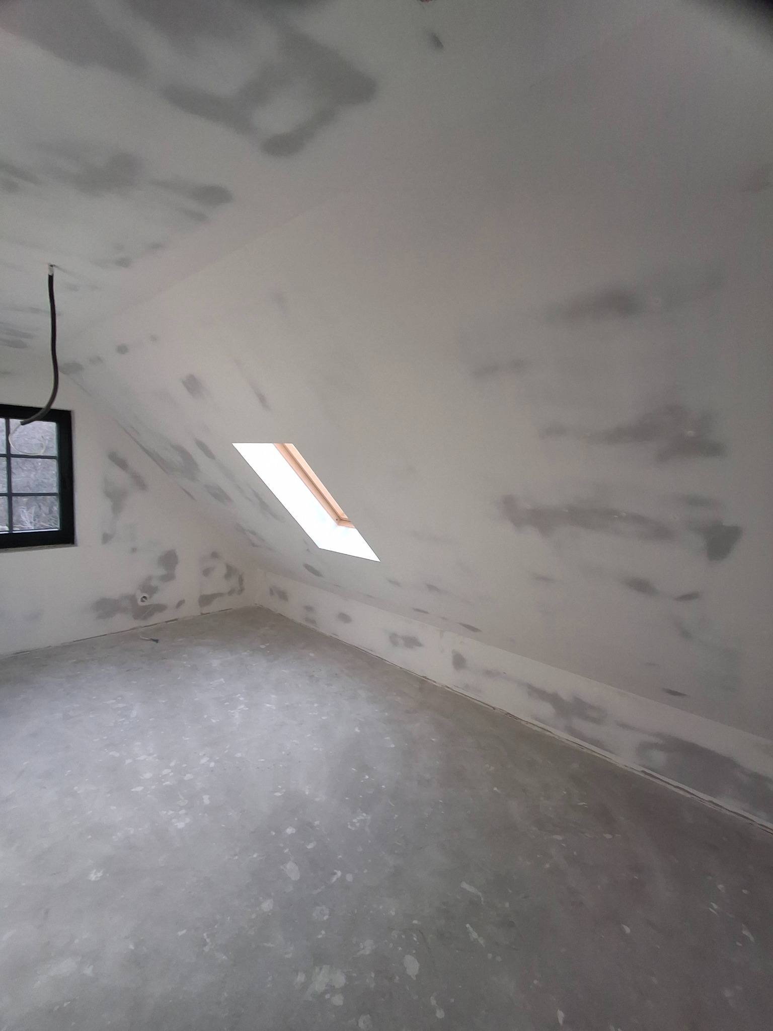 Poddasze w trakcie prac wykończeniowych: surowe ściany i sufit z nałożoną warstwą szpachli, widoczne okno dachowe z drewnianą ramą i czarna rama okienna na bocznej ścianie.