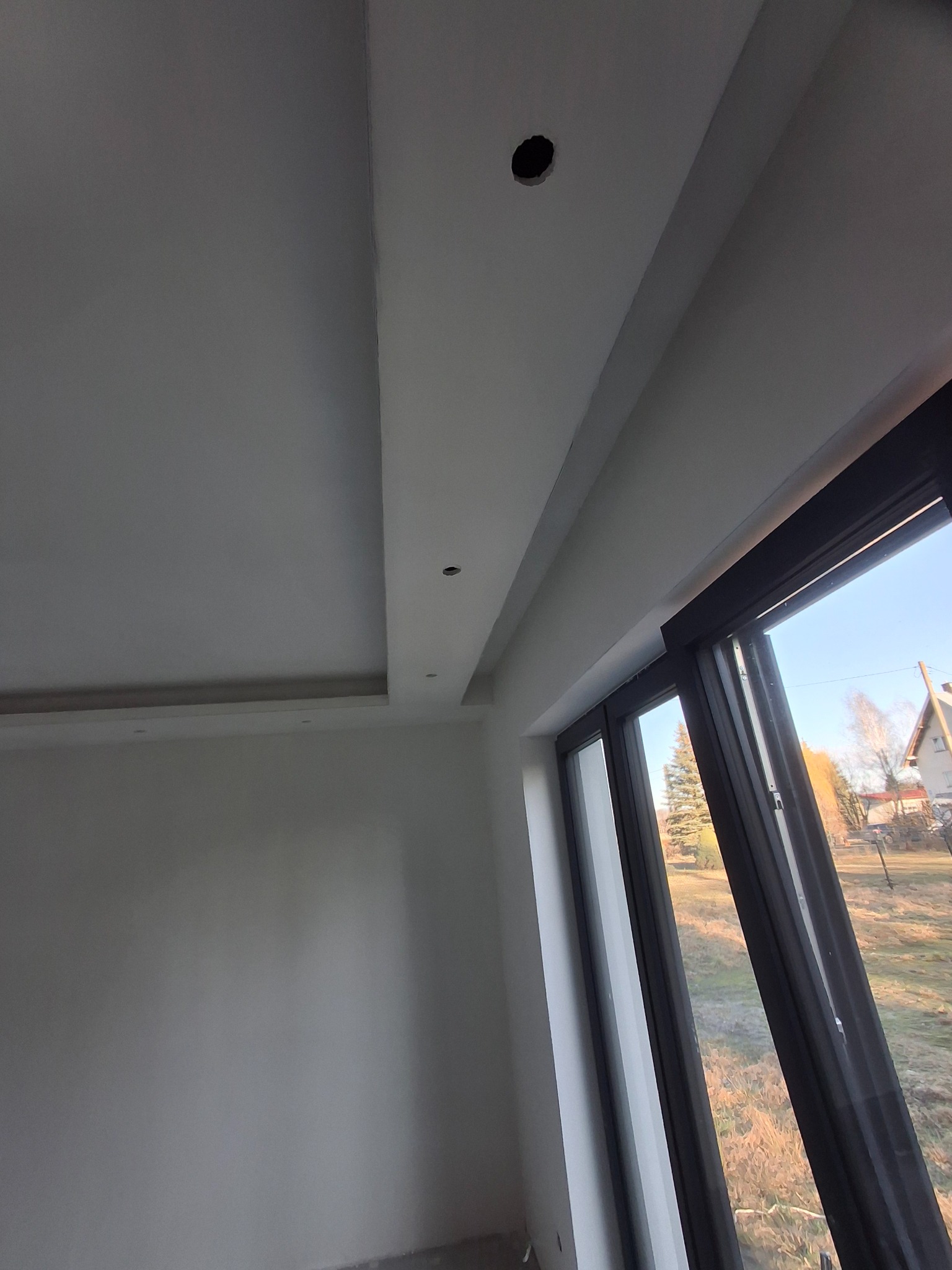 Wnętrze pokoju po gładziowaniu i malowaniu, widoczne otwory na oświetlenie w suficie podwieszanym i duże okno z widokiem na zewnątrz.