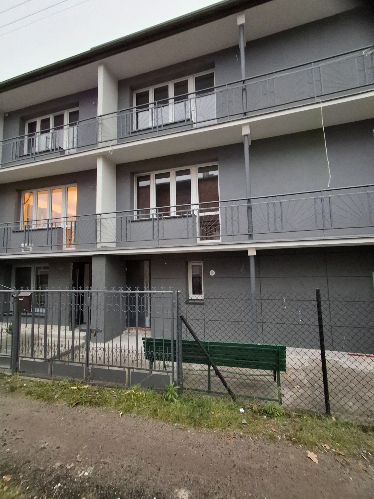 Szary budynek wielorodzinny z balkonami i ozdobnymi balustradami, widziany zza metalowego ogrodzenia. W tle zielona ławka.