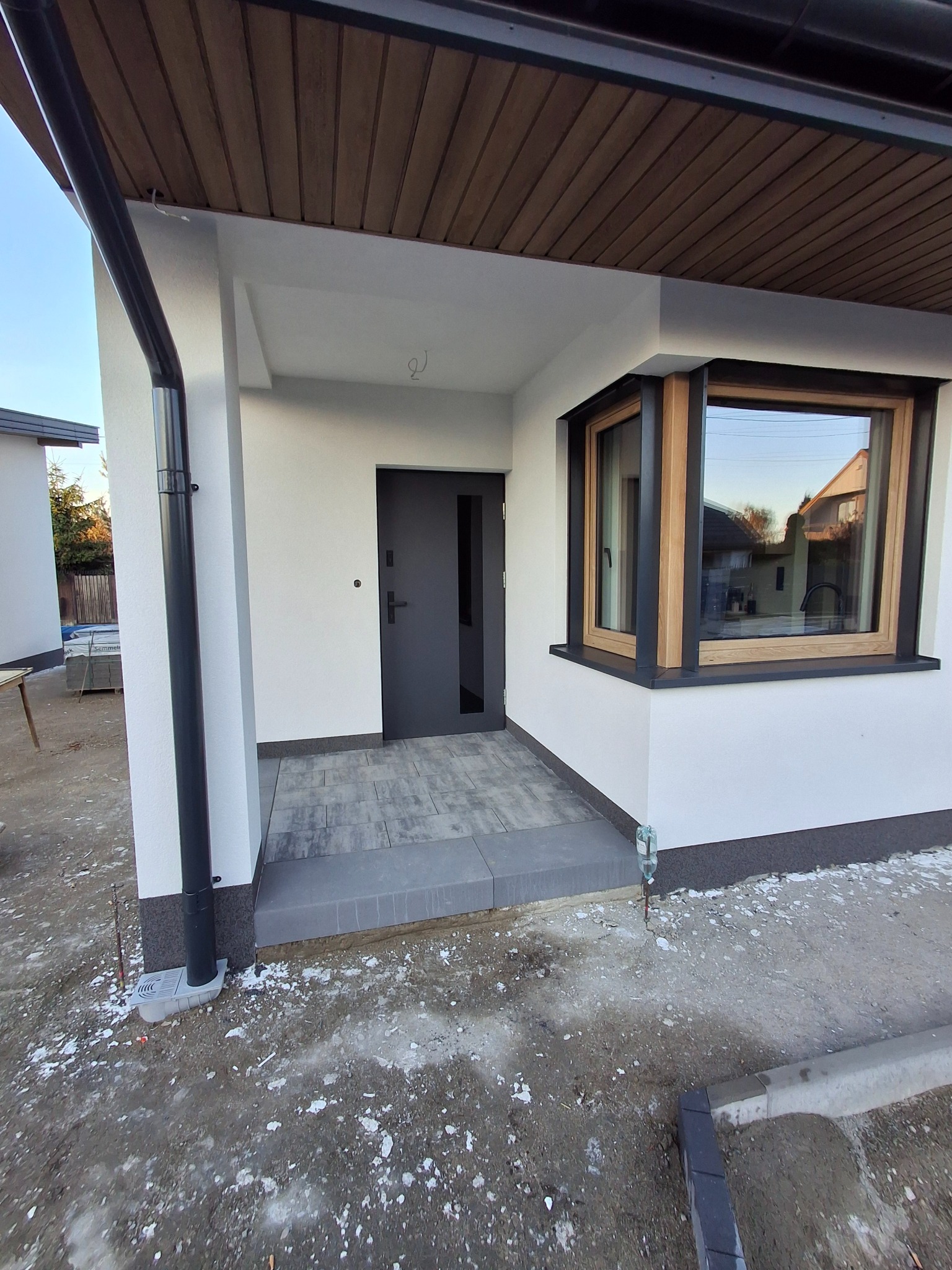 Wykończone wejście do domu z szarą kostką, białą elewacją i drewnianą podbitką. Okno z drewnianą ramą. Widoczny fragment rynny.