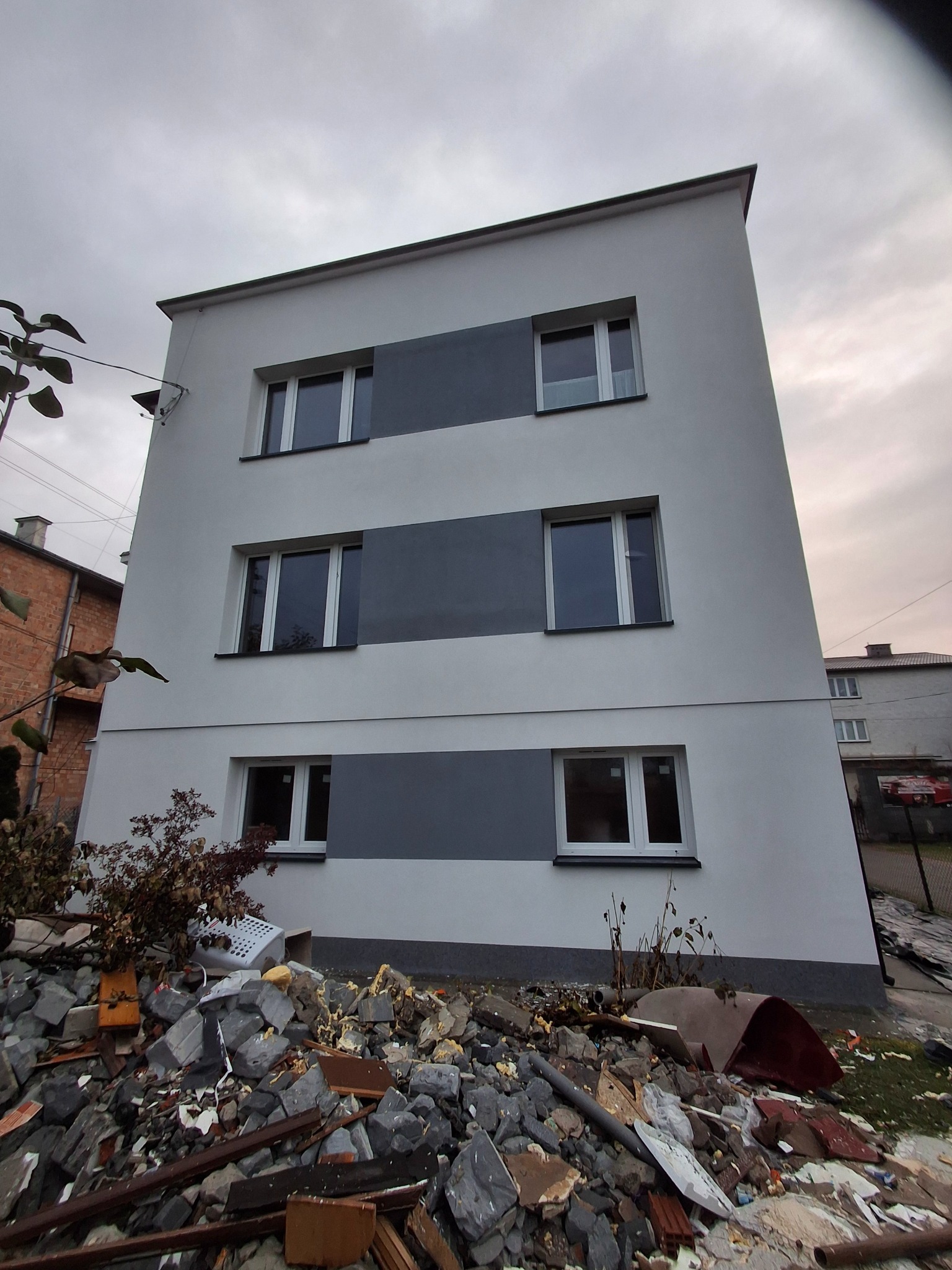 Odnowiona elewacja trzypiętrowego budynku z szarymi pasami, widoczna z dołu, z gruzem budowlanym u podstawy. Świeże malowanie, nowe okna, pochmurne niebo.