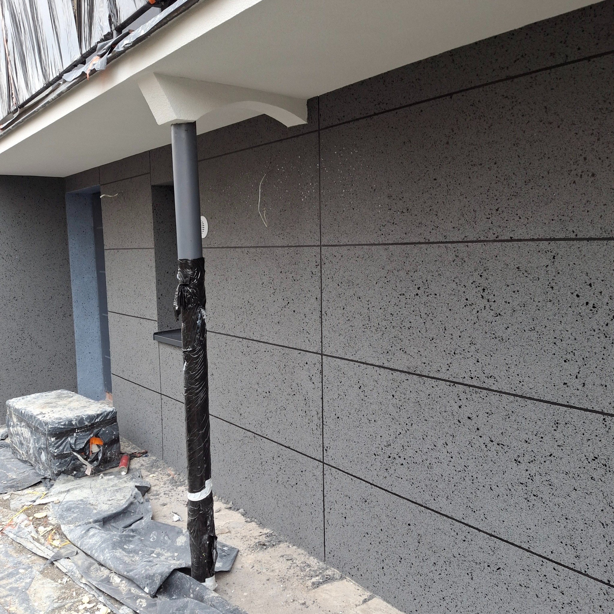 Elewacja budynku wykończona szarymi płytami z ciemnymi punktami, zabezpieczona rura spustowa, widoczne materiały budowlane, etap prac wykończeniowych.