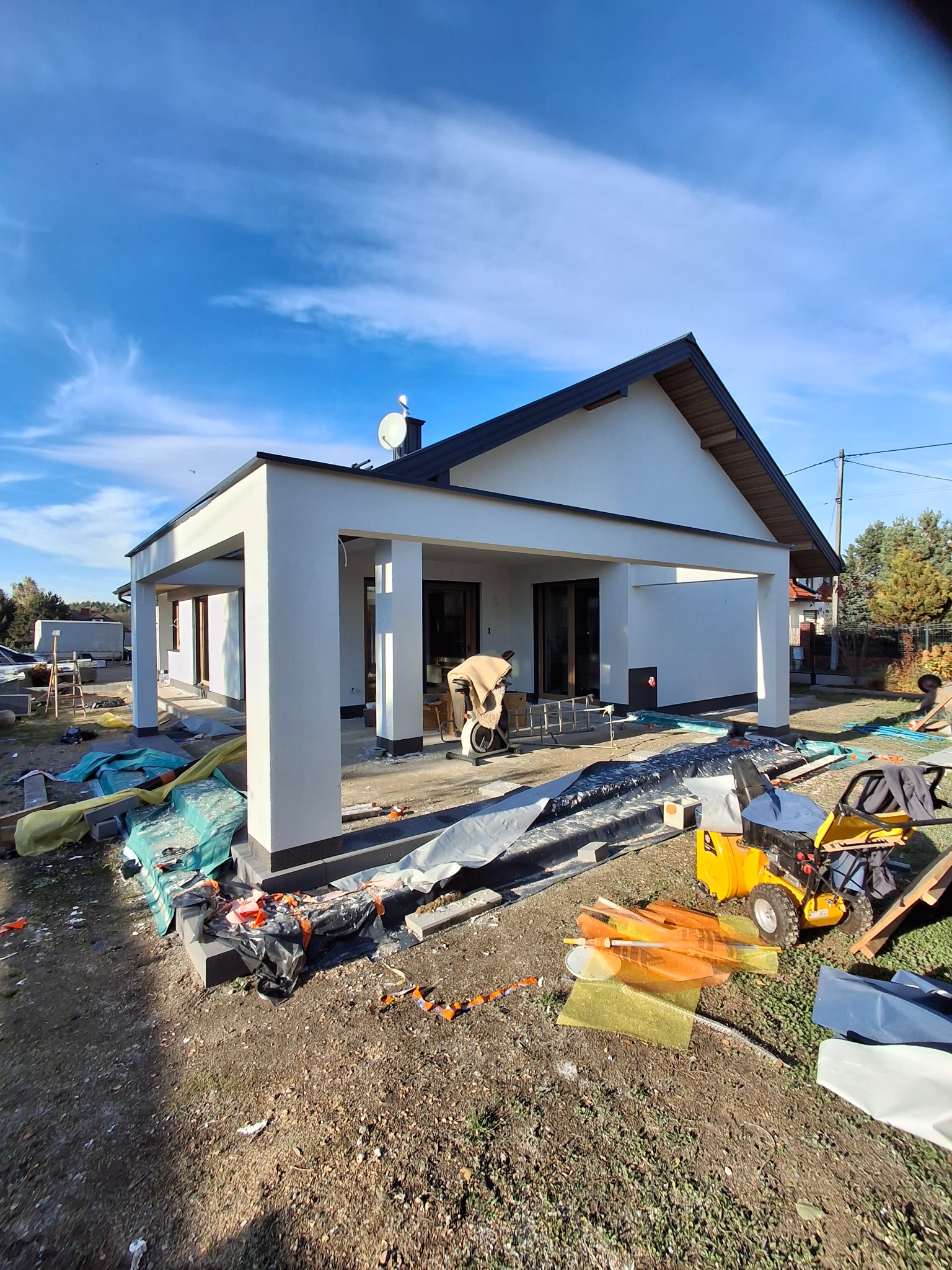 Biały budynek z kolumnami w trakcie wykończenia elewacji, widoczne narzędzia i materiały budowlane, błękitne niebo w tle. Prace wykończeniowe.