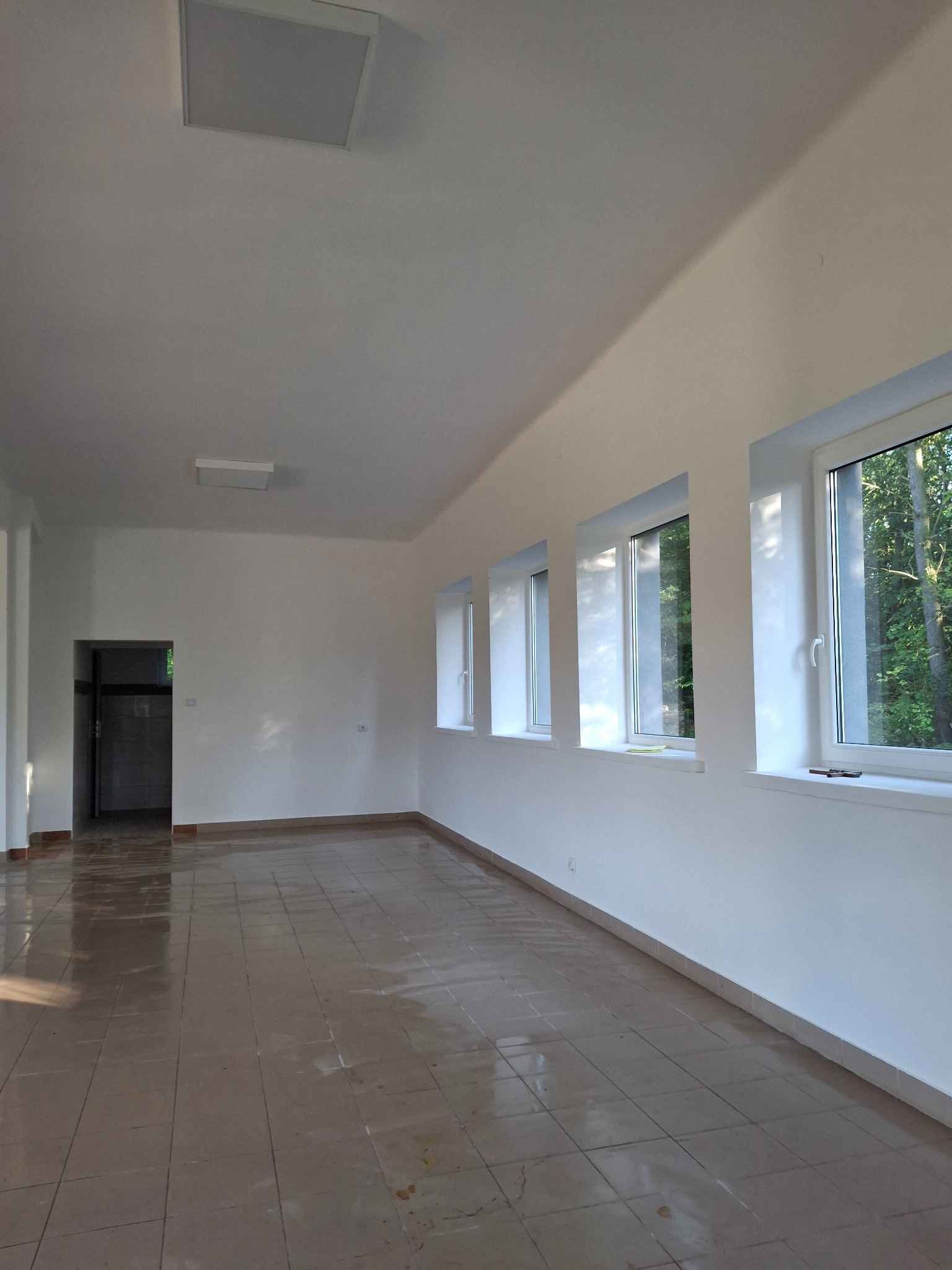 Jasne, przestronne wnętrze po remoncie: białe ściany, brązowa podłoga z płytek, okna z widokiem na zieleń. Widoczne ślady po remoncie na podłodze.