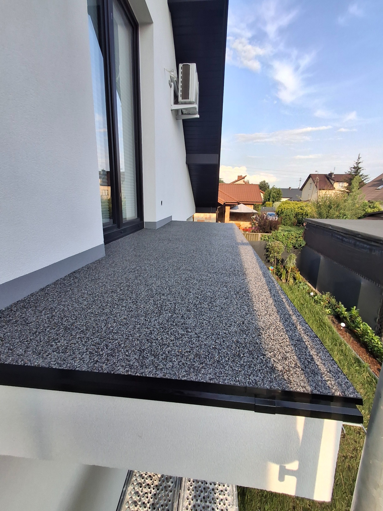 Wykończony balkon z szarą, kamyczkową posadzką i czarnym obramowaniem, widok z góry na tle zieleni i zabudowań. Widoczny fragment elewacji budynku.