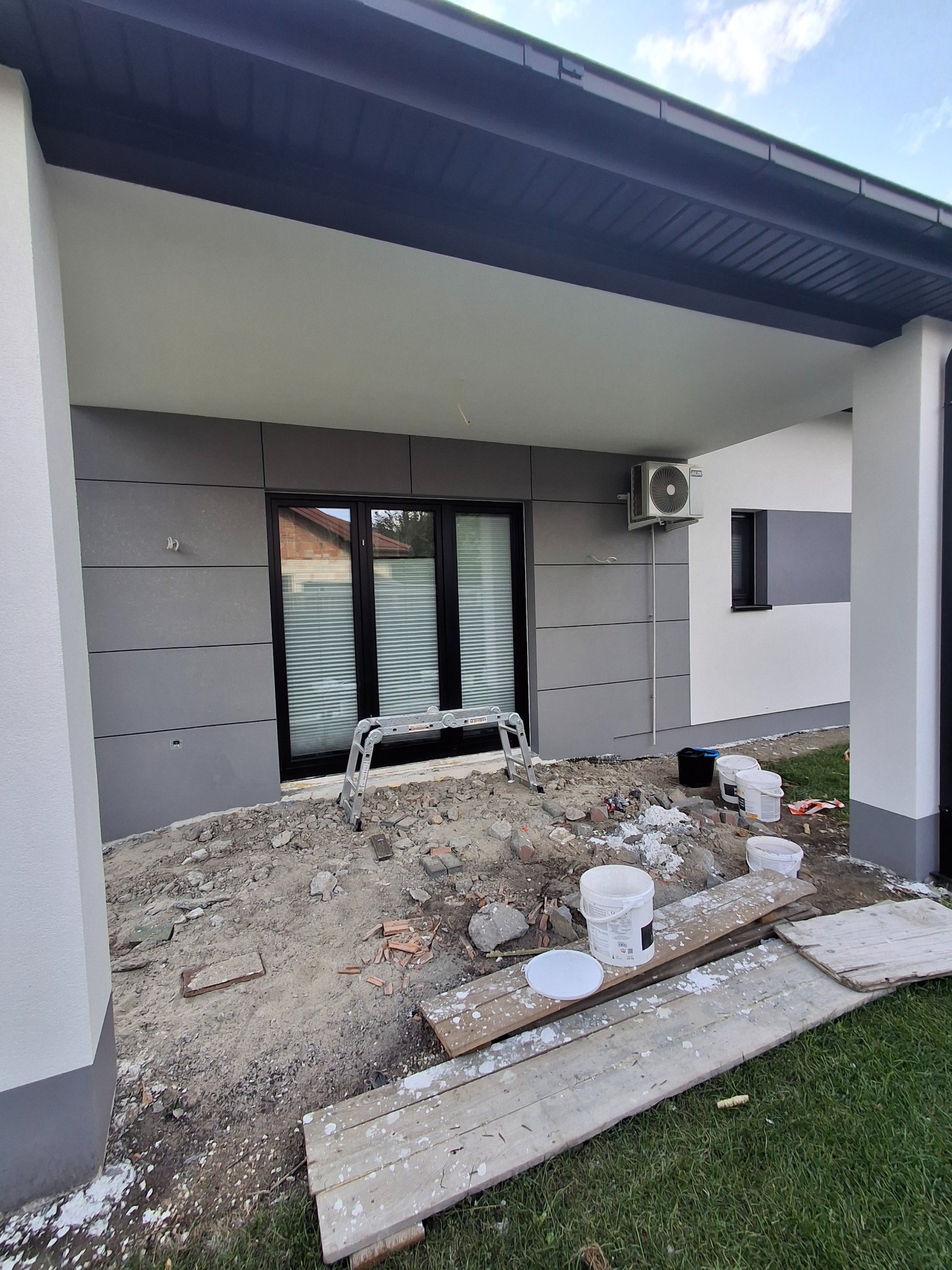 Elewacja domu z szarymi panelami i czarną stolarką okienną. Widoczne przygotowania do prac wykończeniowych, drabina i farby. Częściowo widoczny klimatyzator.