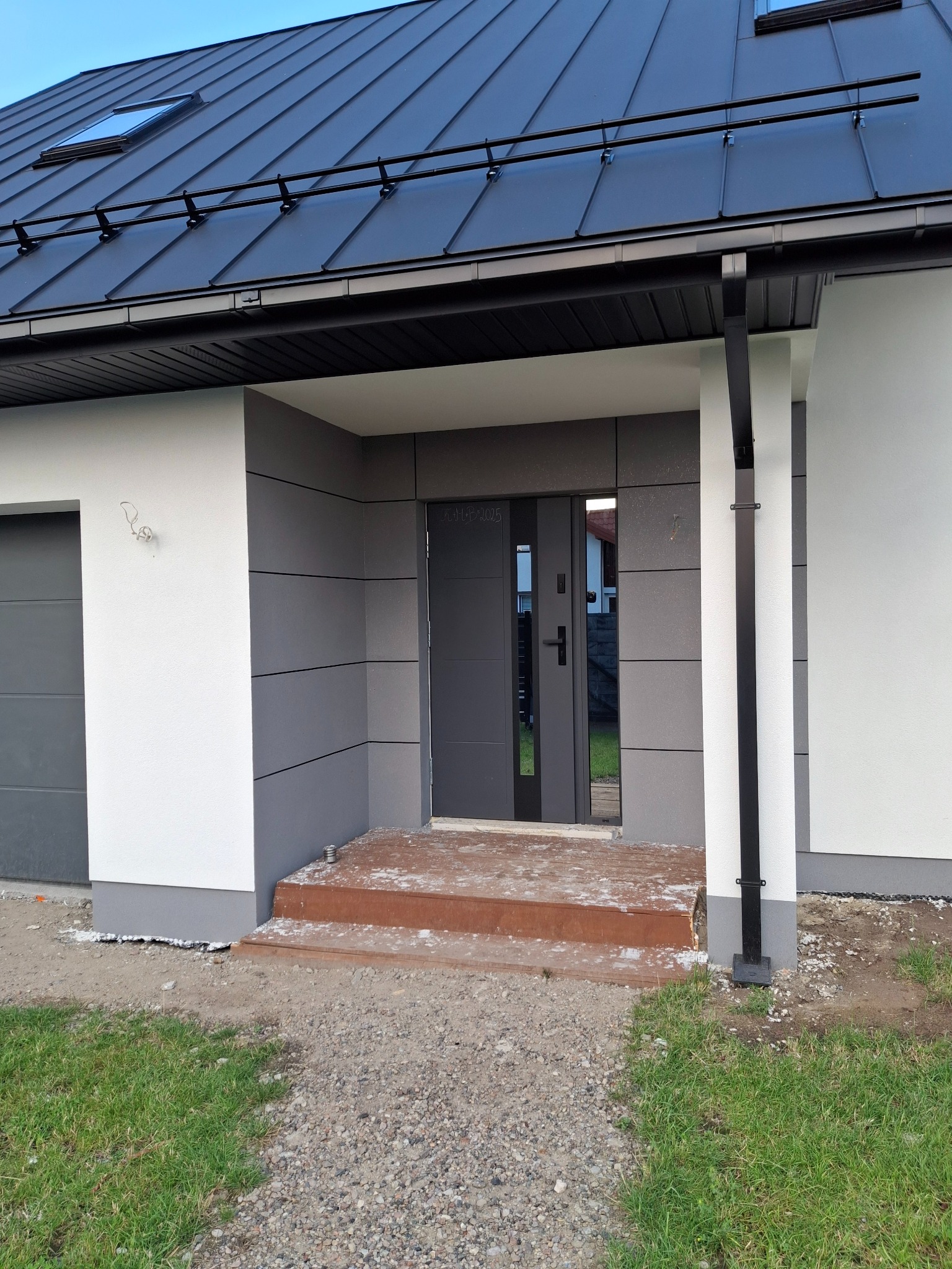 Nowoczesny dom jednorodzinny z grafitową elewacją i ciemnym dachem. Widoczne wejście z drewnianymi schodami i fragment ogrodu. Minimalistyczny design.