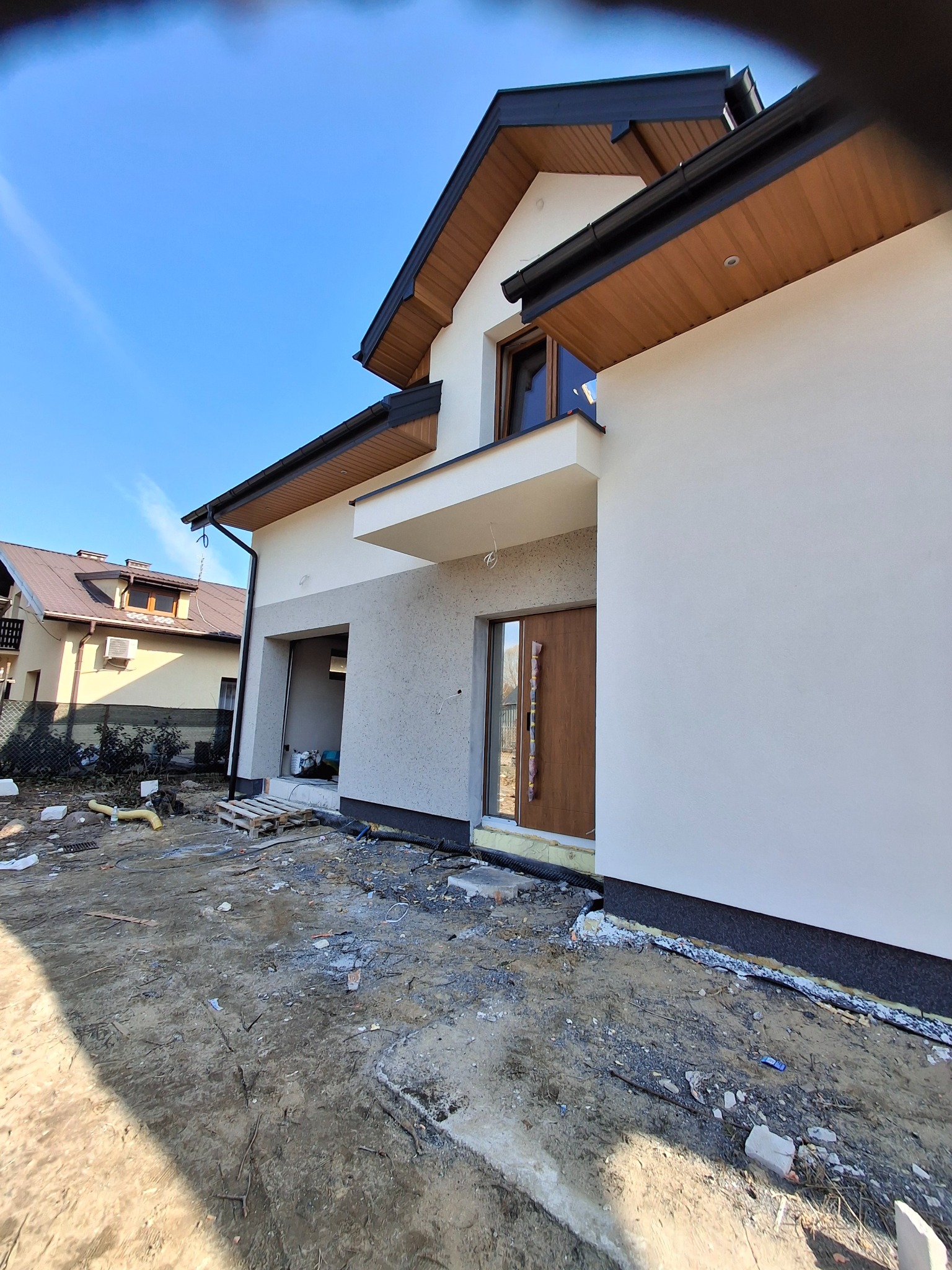 Nowoczesny dom jednorodzinny z jasną elewacją i drewnianymi elementami wykończeniowymi. Widoczne prace wykończeniowe wokół budynku, błękitne niebo w tle.