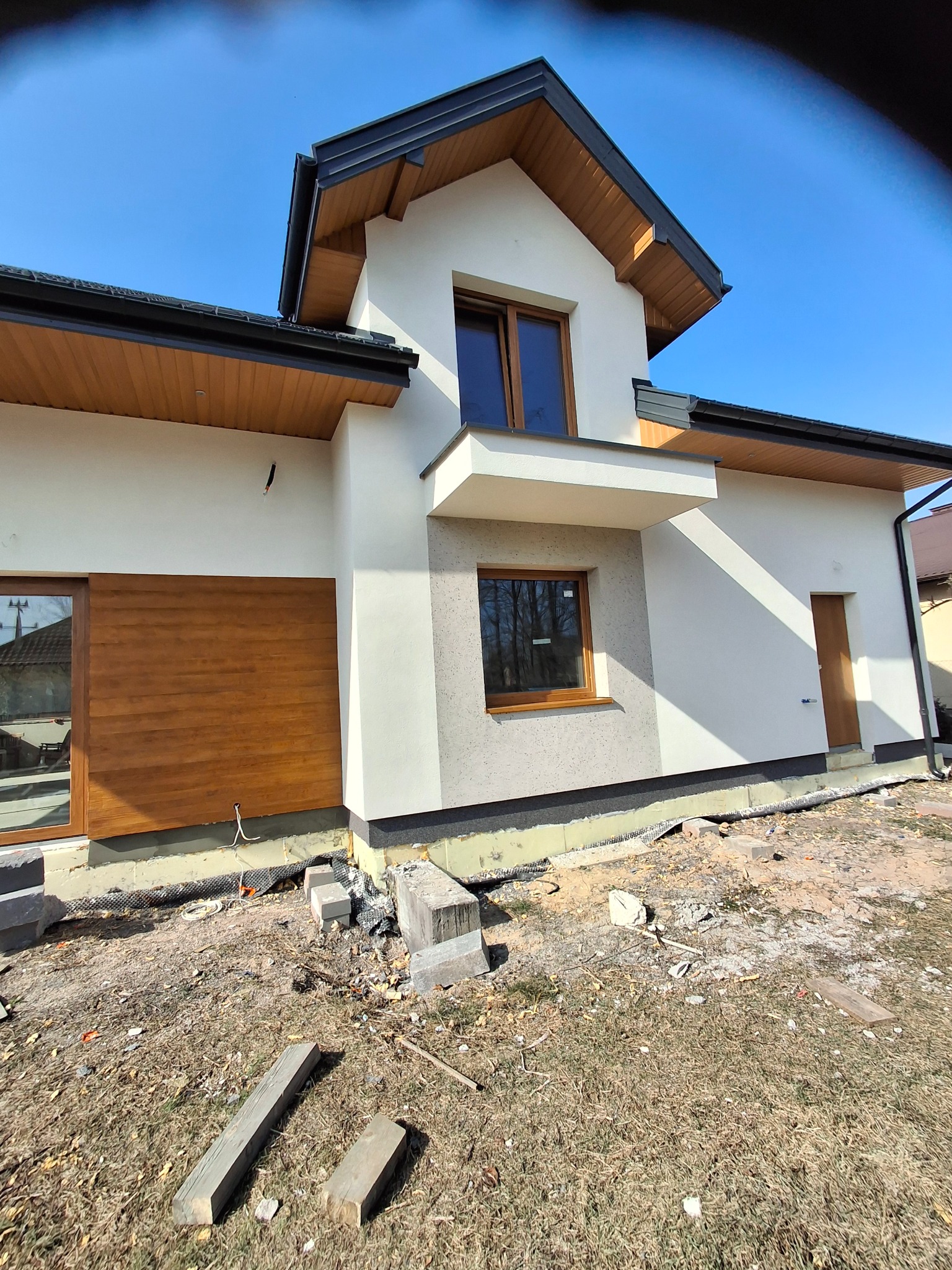 Nowoczesny dom jednorodzinny z elementami drewnianymi na elewacji, w trakcie prac wykończeniowych. Widoczne okna, drzwi i fragment elewacji z fakturą.