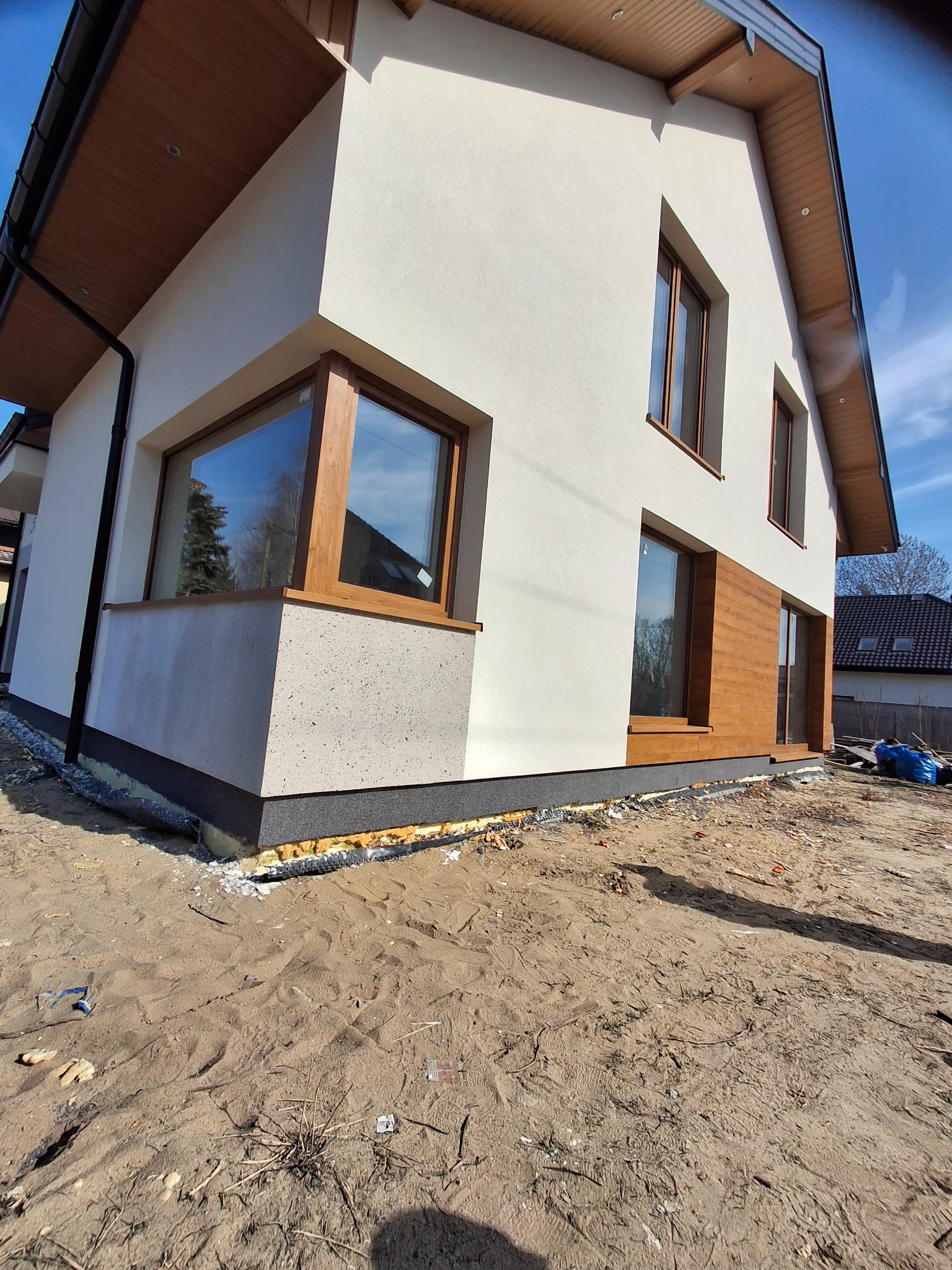 Nowoczesny dom jednorodzinny z elewacją w odcieniach bieli i drewna, widok z zewnątrz, teren wokół domu w trakcie prac wykończeniowych.