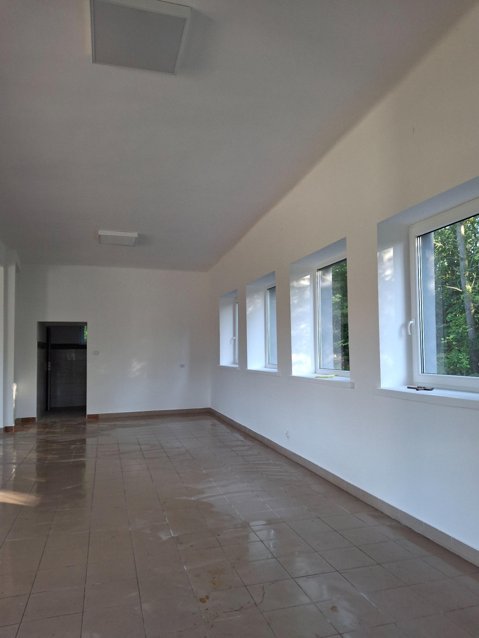 Wnętrze po remoncie: białe ściany, brązowa podłoga z płytek, okna z widokiem na zieleń. Widoczne detale wykończeniowe i oświetlenie sufitowe.