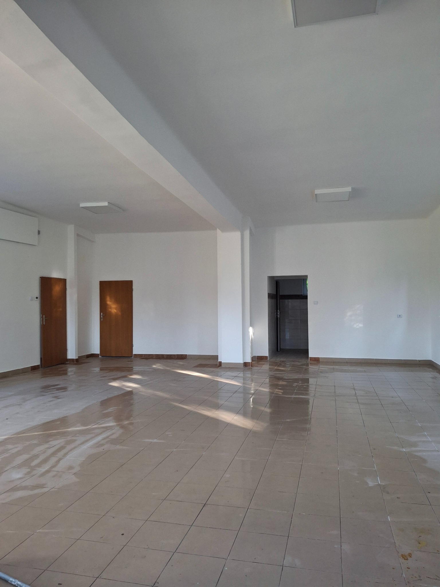 Jasne, przestronne wnętrze po remoncie: białe ściany, brązowe drzwi i podłoga z płytek. Widoczne ślady po pracach wykończeniowych, wpadające promienie słoneczne.