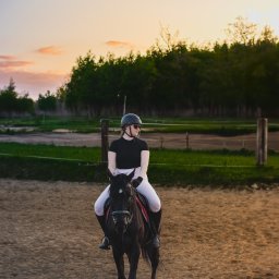 Bartosz Błażek Fotografia - Kobieta w czarnym stroju jeździeckim i kasku na czarnym koniu na piaszczystym padoku, w tle drzewa i zachodzące słońce.