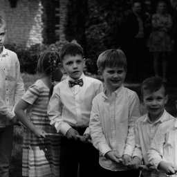 Bartosz Błażek Fotografia - Czarno-białe zdjęcie grupy dzieci, w tym jeden chłopiec w muszce i z medalikiem, trzymających linę podczas zabawy na zewnątrz, z dorosłymi w tle.