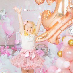 Bartosz Błażek Fotografia - Uśmiechnięta blond dziewczynka w różowej tiulowej spódniczce i bluzce z łabędziami stoi na tle pastelowych balonów i dekoracji urodzinowych, z rękami uniesionymi do góry w geście radości, obok...