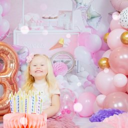 Bartosz Błażek Fotografia - Uśmiechnięta blond dziewczynka w różowej tiulowej spódniczce siedzi na tle dekoracji urodzinowych w odcieniach różu i złota, w tym balon w kształcie cyfry 3, tort z papierowymi świeczkami i wózek...