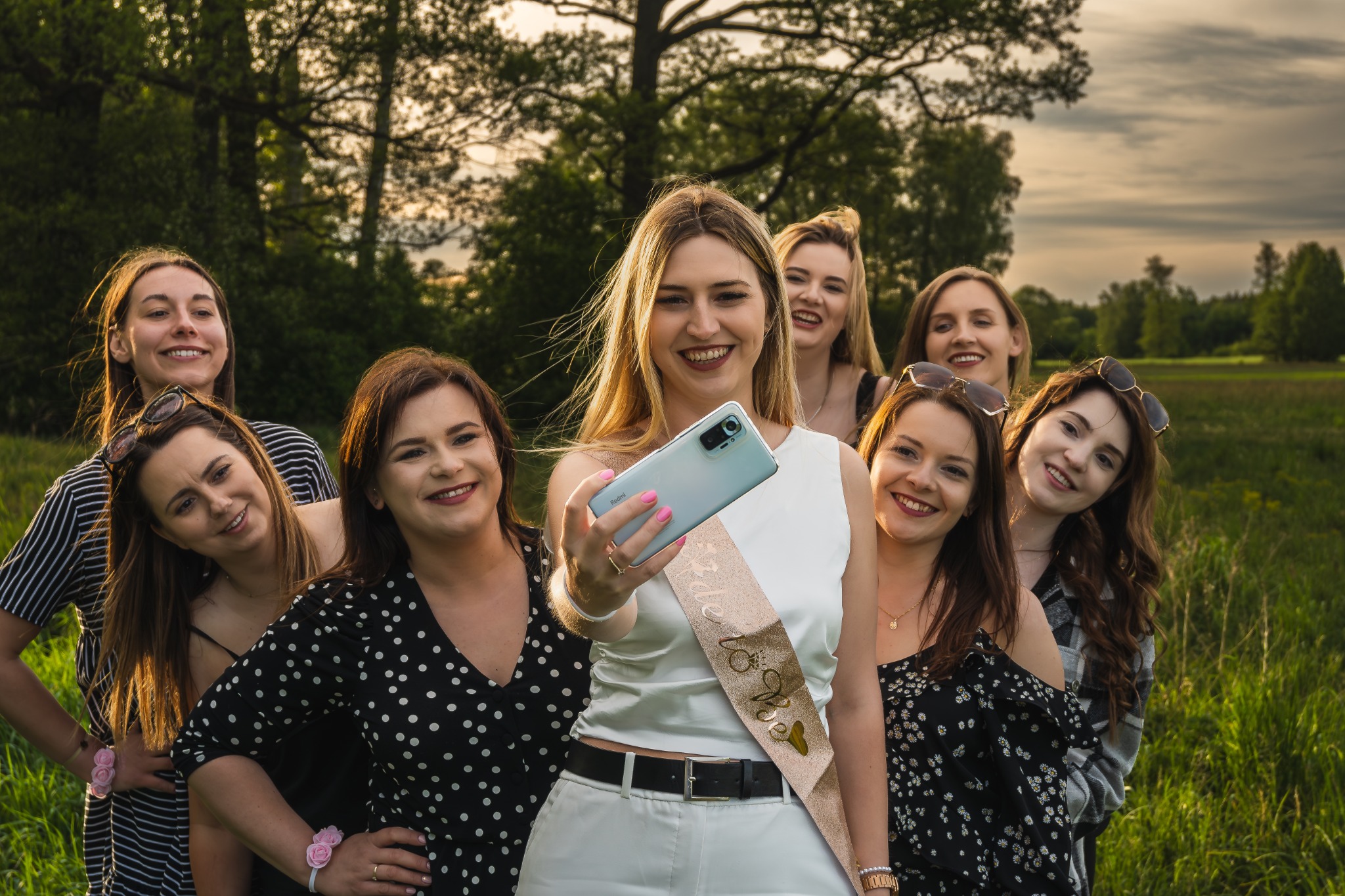 Panna młoda z druhnami robi selfie w plenerze, panna młoda w białym stroju i szarfie 'Bride to be', uśmiechnięte kobiety pozują na tle zielonej łąki i drzew.
