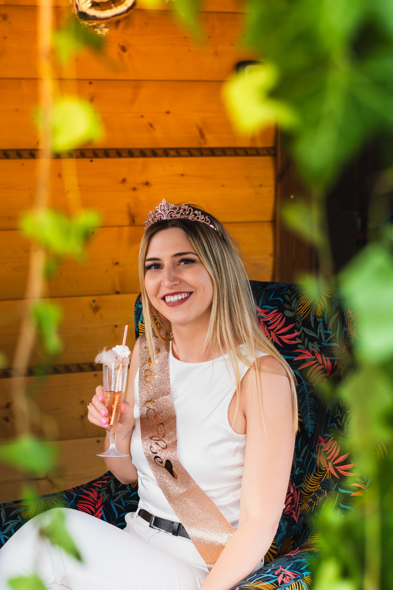 Uśmiechnięta blondynka w tiarze i szarfie 'Bride to Be' siedzi w fotelu z tropikalnym wzorem, trzymając kieliszek z różowym napojem i dekoracją z waty cukrowej, na tle drewnianej ściany i zielonych...