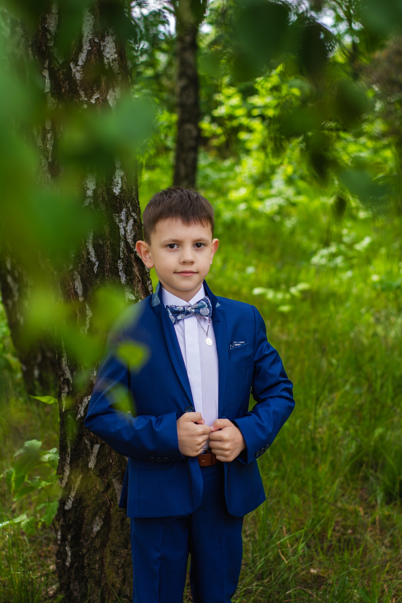 Chłopiec w granatowym garniturze i muszce, pozujący przy brzozie w leśnym otoczeniu.