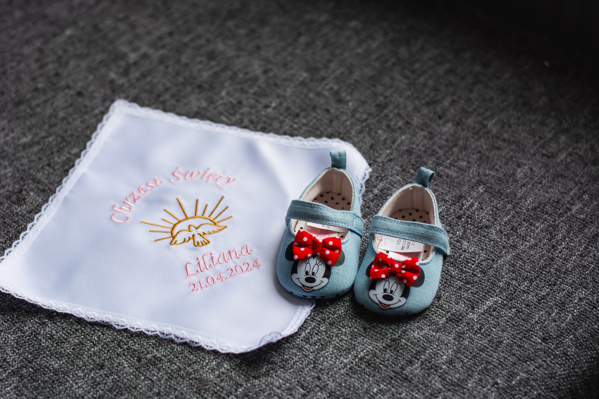 Biała haftowana chusteczka z napisem 'Chrzest Święty' i datą 21.04.2024 obok pary dżinsowych bucików niemowlęcych z motywem Myszki Miki i czerwoną kokardką, ułożonych na szarym tle.