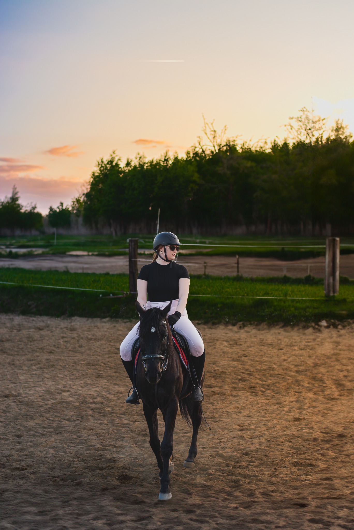 Kobieta w czarnym stroju jeździeckim i kasku na czarnym koniu na piaszczystym padoku, w tle drzewa i zachodzące słońce.