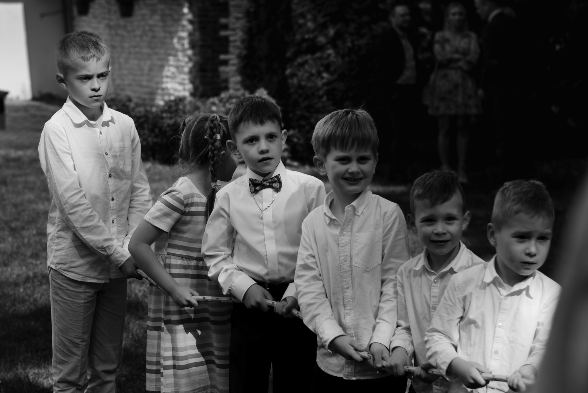 Czarno-białe zdjęcie grupy dzieci, w tym jeden chłopiec w muszce i z medalikiem, trzymających linę podczas zabawy na zewnątrz, z dorosłymi w tle.