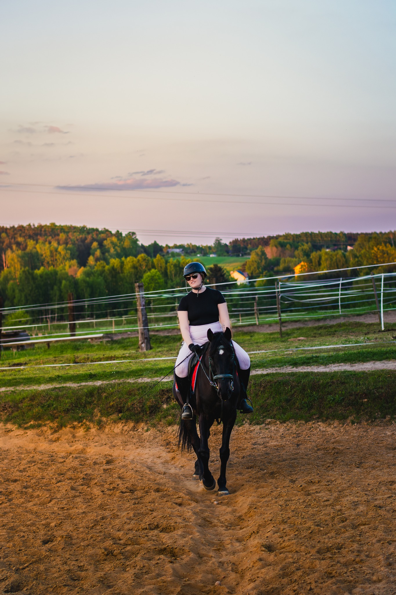 Kobieta w kasku i okularach przeciwsłonecznych jeździ na czarnym koniu po piaszczystym terenie, w tle widoczne drzewa i zachodzące słońce.