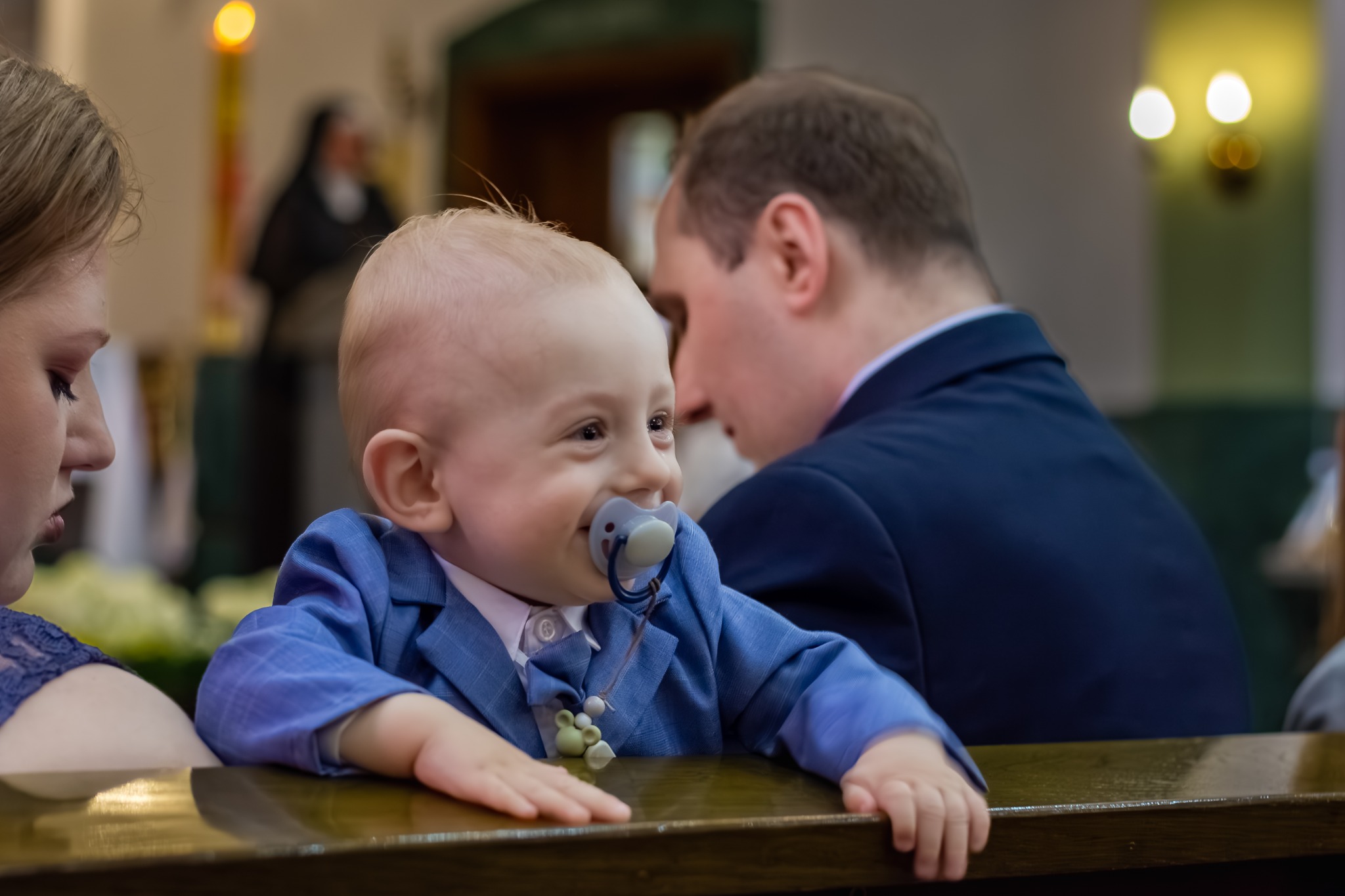 Uśmiechnięty niemowlak w niebieskim garniturze z smoczkiem, oparty o drewniany stół podczas uroczystości w kościele, w tle zamazane postacie.