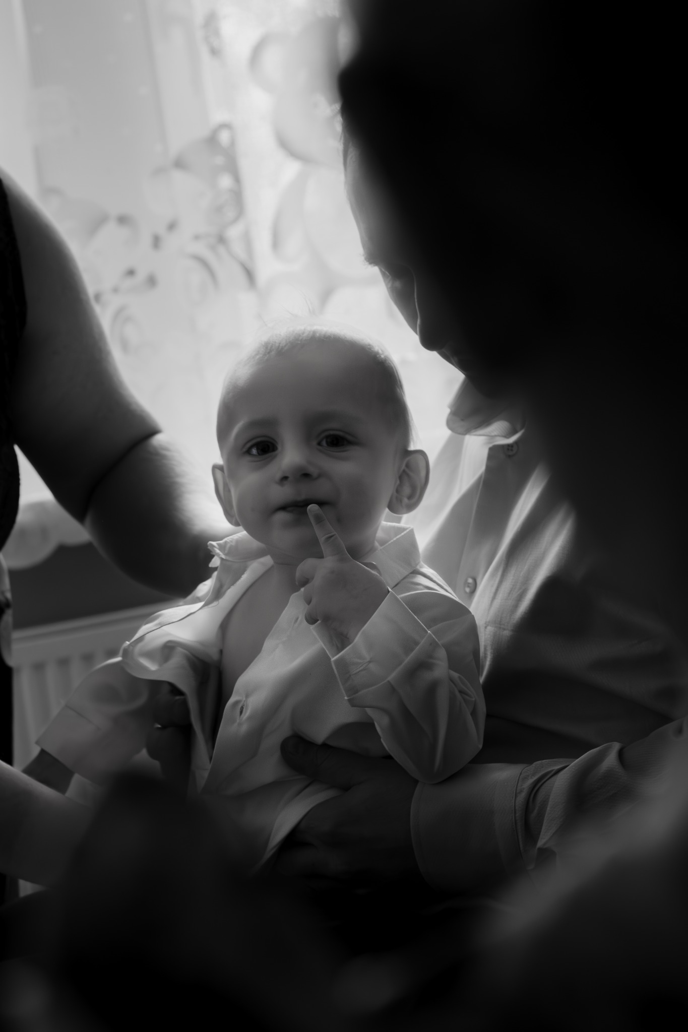 Czarno-białe zdjęcie niemowlęcia w białej koszuli, trzymanego na rękach, z palcem przy ustach i spojrzeniem skierowanym w obiektyw, uchwycone w intymnej atmosferze w pobliżu okna.