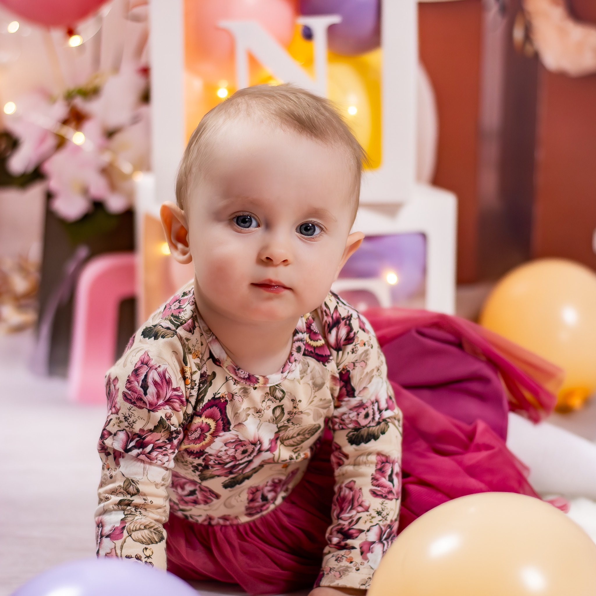 Urocze dziecko w kwiecistej bluzce i różowej spódniczce, siedzące na podłodze z balonami i dekoracjami, w tle białe litery z lampkami.