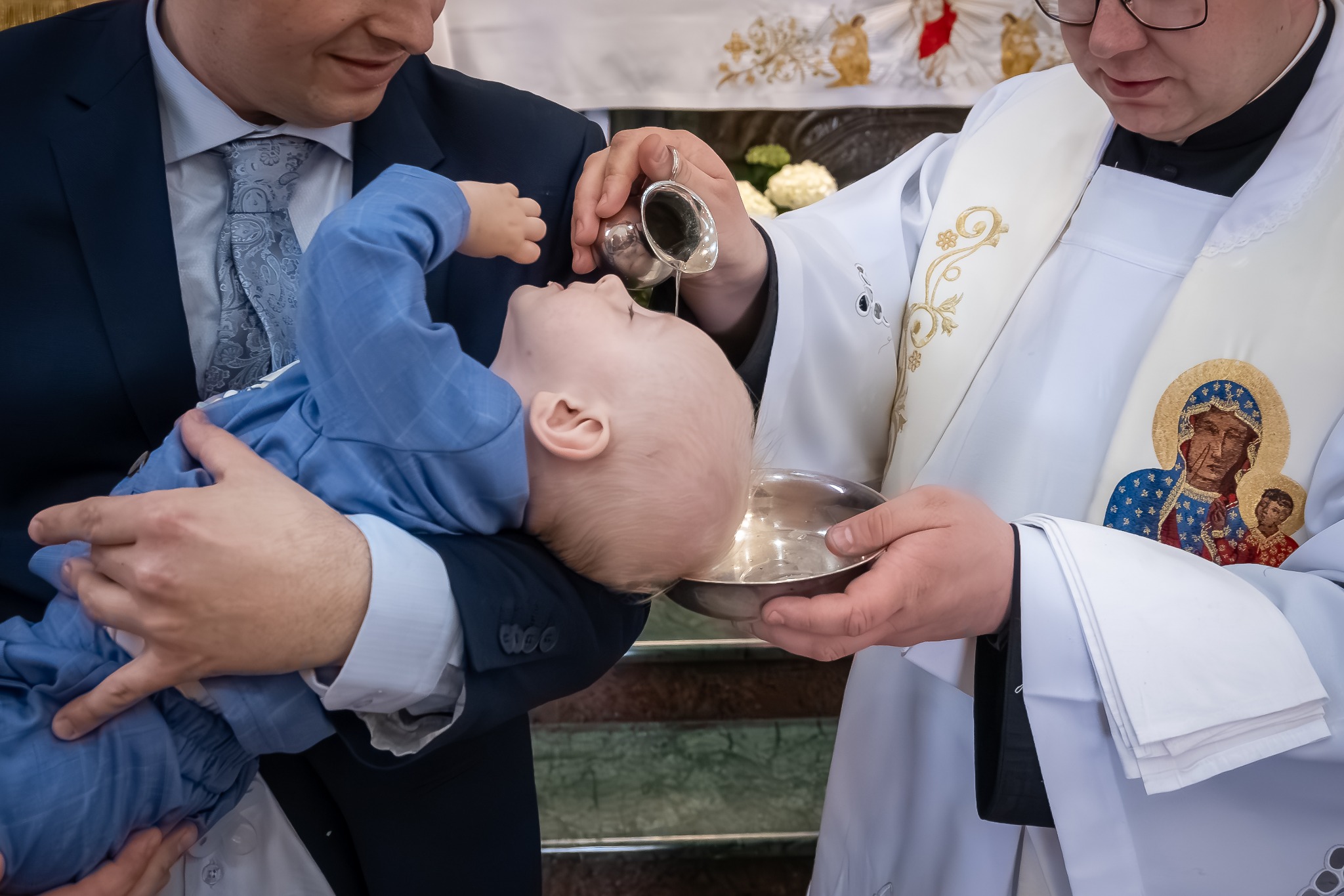 Ksiądz polewa wodą głowę niemowlęcia trzymanego przez mężczyznę podczas ceremonii chrztu.