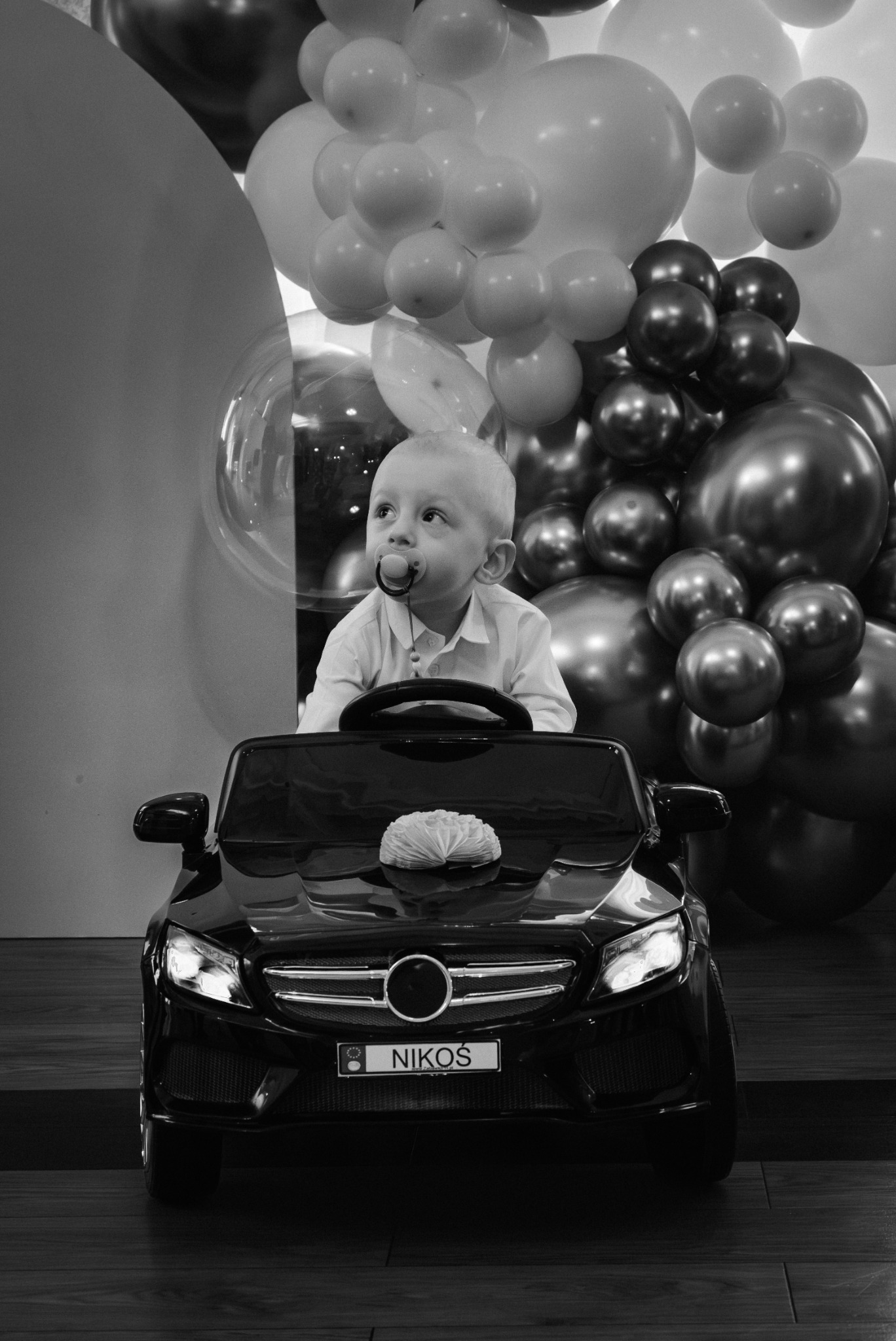 Czarno-białe zdjęcie małego chłopca z smoczkiem w ustach, siedzącego za kierownicą czarnego, zabawkowego samochodu na tle balonów w różnych odcieniach szarości, z tablicą rejestracyjną z napisem...